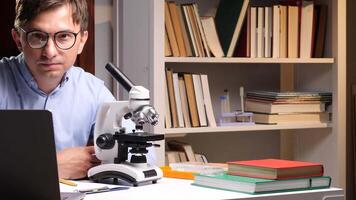 Close-up portrait male scientist sitting at desk using laptop and microscope, conducting research in modern laboratory with books and equipment, focused on scientific analysis and discovery video