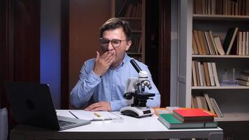 student, scientist at desk with microscope tired and yawning. Tired man yawning. Overworked scientist needs rest. Desk with microscope and laptop video