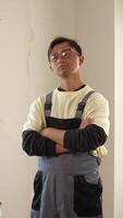 Vertical portrait of a serious construction worker with glasses and overalls, crossing arms confidently in front of a damaged wall at a renovation site video