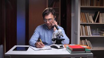 Scientist working late at night in laboratory taking notes while talking on phone video
