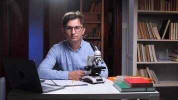 portrait of a Young scientist sitting at desk with microscope, laptop, and books, working late in a laboratory or home office, conducting research or analysis video