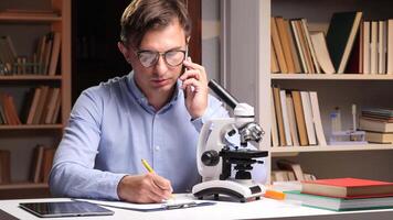 Close-up portrait scientist taking notes while talking on the phone, with a microscope, books, and tablet in a laboratory setting, conducting research and analysis video