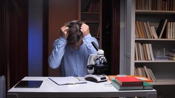 Young scientist covering face with hands, feeling overwhelmed and fatigued from long hours in the laboratory, surrounded by a microscope, books, and a tablet on the desk video