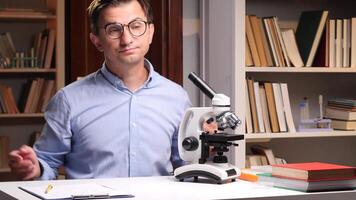 Close-up portrait a confused scientist is shrugging his shoulders while sitting at a desk with a microscope and books in a laboratory, unsure of the results of his experiment video