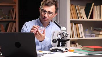 Close-up portrait scientist examining a petri dish sample under a microscope in a lab filled with books and a laptop, advancing research in biology and chemistry video