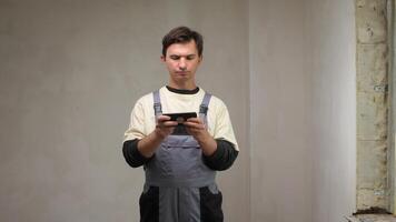Construction worker wearing overalls using a smartphone in an unfinished room, likely checking blueprints, sending messages, or managing project details video