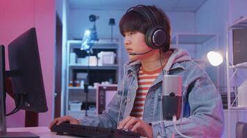 Young Asian teenage gamer is excited playing online games on gaming computer with keyboard and headset, cyber virtual technology, esports entertainment in dark room, fun teenage streaming lifestyle. photo