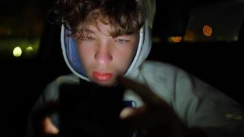 Adolescente masculino pasajero dentro coche durante Noche, inmerso en teléfono inteligente pantalla, creando suave ciudad luces bokeh en oscurecido fondo, capturar contemporáneo digital estilo de vida momento video