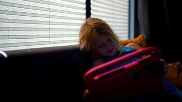 Blonde girl using tablet while relaxing on couch at home with soft lighting and blinds behind, filmed with vintage lens video