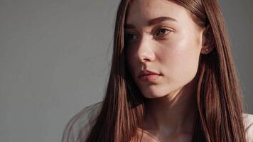Soft sunlight filtering through studio space, highlighting contemplative young woman with flowing brown hair, posing elegantly against neutral palette backdrop video