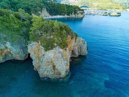 Cliff with wild vegetation above the waves of the Adriatic Sea against the backdrop of the city of Budva and the Montenegrin mountains photo