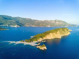 Island of St. Nicholas with vegetation on shores in the Adriatic Sea against the backdrop of coastal towns, the Montenegrin mountains and clear sky photo