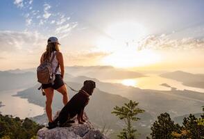 A sporty girl with a backpack stands on the edge of a mountain with a Rottweiler dogSporty girl with backpack and Rottweiler on mountain edge photo
