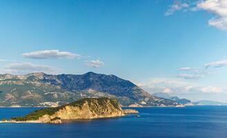 Island of St. Nicholas with vegetation on shores in the Adriatic Sea against the backdrop of coastal towns, the Montenegrin mountains and clear skyIsland of St Nicholas with Coastal Towns photo