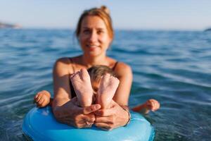 A young mother shows the legs and arms of her baby, who is sitting in an inflatable ring in the seaYoung mother baby legs arms inflatable ring sea photo