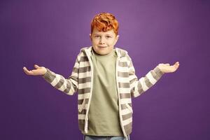 Puzzled and ignorant little red-haired boy shrugs his shoulders, symbolizing lack of knowledge, showing illiteracy wearing in casual clothes standing against violet isolated background photo