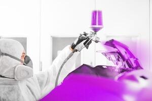 Spray painter worker in protective glove with airbrush pulverizer painting car element in white paint chamber. photo
