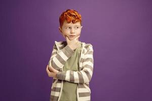 Pensive Red Headed Little Boy Around 7-8 years Puzzled Standing Keeping Hand to Cheek with Thoughtful Face Expression Looking Aside, Wearing in Casual Clothes Standing on Isolated Violet Background photo