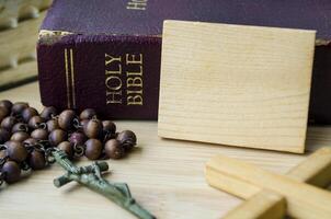 Holy Bible and Holy Rosary placed beside wooden block with customizable space for text or message sharing. Christianity concept photo