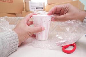 Hands unboxing or packing a ceramic mug with bubble wrap, showing fragile item care, shipping, surrounded by other delivery packages. photo