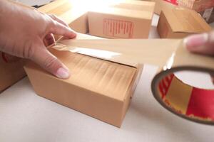 Hands sealing cardboard box with brown packing tape for shipping, surrounded by other delivery packages. photo