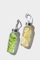 Top view two glass bottles, water drink detox with lemon, cucumber at sunlight on white background, copy space. Infused water with slices fruit or vegetable. Aesthetic photo with fresh drink