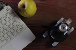 There is a microscope and a laptop on the wooden table. The emphasis is on the apple. photo