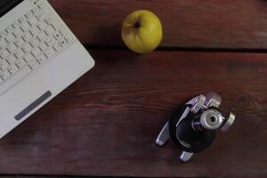 There is a microscope, a laptop and a green apple on the wooden table. photo