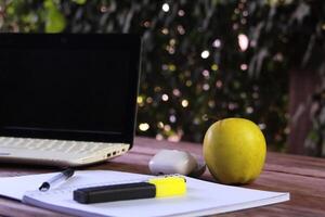 A laptop lying on a wooden surface outdoors. Next to the laptop is a yellow apple, as well as an open notebook with a pen and a yellow marker. photo
