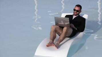 Concept remote work, freedom and digital lifestyle. Young freelancer or entrepreneur in blazer and swim trunks working on a laptop by the pool. video
