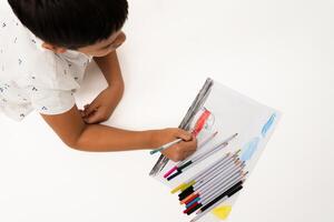 Young boy drawing on white paper with colored pencils, viewed from above on an isolated white background. Suitable for education, creativity, childhood, and art-related concepts. photo