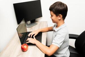Young boy in a gray polo shirt sits at a wooden desk using a laptop, with an apple and a monitor nearby. Suitable for illustrating remote learning, home study, or technology use by children. photo