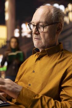 Retired old person with spectacles enjoys reading a book at the library, being happy and enjoying recreation. Aged man pensioner with book reading hobby, retirement leisure activity in quiet space. photo