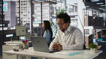 Black man employee joins a telecommuting session at his office desk, multitasking between project management and business strategies. Productivity and collaboration across departments. Camera B. photo