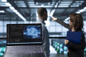 Laptop in front of data center technicians in blurry background deploying AI driven management tools, inspecting server clusters, updating network topologies, validating storage integrity photo