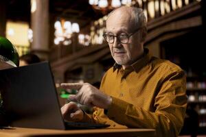 Elderly wise college student interacting with professor at webinar on laptop, studying in a library and combining modern telecommunication with classic academic research. Seminar lesson. photo