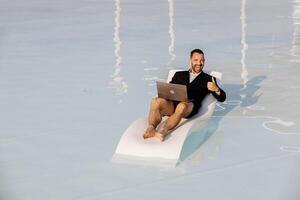 A man in a jacket, swimming trunks and glasses, relaxing by the pool, works intently on a laptop. The concept of remote work and modern business lifestyle photo