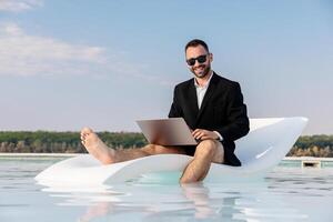 Young freelancer or entrepreneur in blazer and swim trunks working on a laptop by the pool. Concept remote work, freedom and digital lifestyle. photo