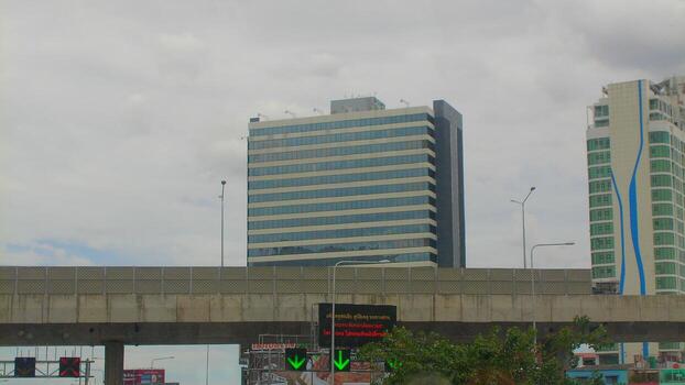 Concrete elevated highway with city buildings visible beyond, captured from street level under a cloudy Bangkok sky, emphasizing transport infrastructure. photo