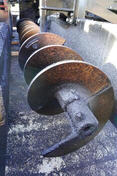 A close-up view of a metal auger with several spiral blades, coated in a dark, gritty substance. The auger is positioned in a machinery setting, showcasing rust and texture. photo