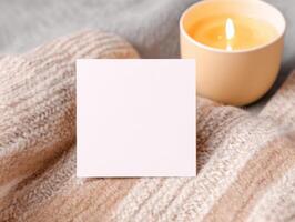 Cozy setting with blank square sticker mockup on soft fabric beside lit candle photo