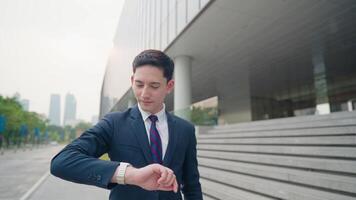 Young mix race light skin businessman in smart attire walking to work on the urban streetside with modern building exterior behind him, confident working person, business owner looking at time video