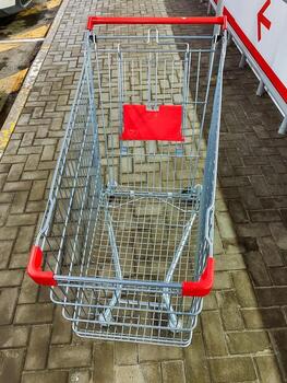 There is a shopping cart that features a vibrant red handle, and it is currently placed on a textured brick sidewalk, creating a striking visual contrast between the two surfaces photo