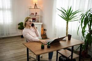 Smiling woman engaged in a phone conversation while working from a cozy home office with plants and natural light photo
