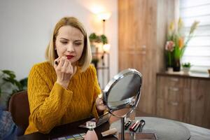 Young woman applying lipstick while seated at a stylish vanity in a cozy modern room with warm lighting photo