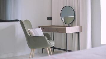 un minimalista vendaje zona presentando un pulcro de madera consola mesa con un redondo espejo, complementado por un felpa aceituna verde silla y un suave almohada, creando un tranquilo esquina en un moderno dormitorio. video