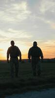 Ukrainian soldiers patrol rural path at sunset. Their silhouettes move calmly as orange light glows in the distance behind fields and trees. video