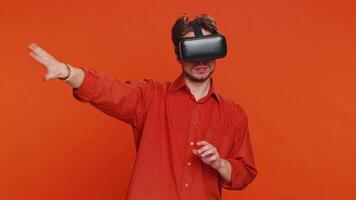 Excited happy man using headset helmet app to play simulation game. Watching virtual reality 3D 360 imagination. Young guy in VR goggles isolated on orange background indoors. Future technology video