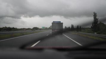 A truck is traveling on a highway surrounded by greenery and trees, with overcast skies and rain creating a moody atmosphere video