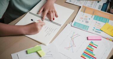 Handheld top view shot, A woman is taking notes in a notebook while sitting at a desk filled with business charts and sticky notes. video
